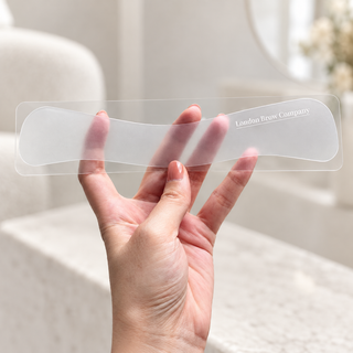 A hand holds a translucent eyebrow-wrap with the printed brand name 'London Brow Company' visible on it, against a softly blurred indoor background.