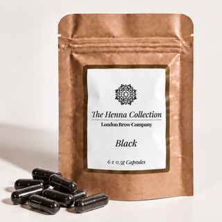 A stand-up kraft paper pouch labeled 'The Henna Collection London Brow Company Black' with several black capsules placed in front on a light neutral background.