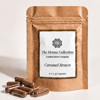 A stand-up kraft paper pouch labeled 'The Henna Collection London Brow Company Caramel Brown' with several brown capsules placed in front on a light neutral background.