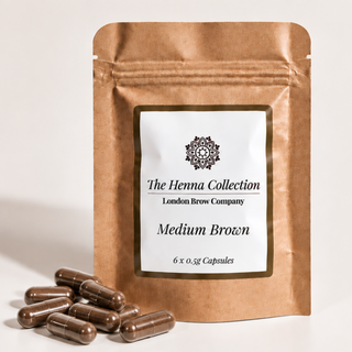 A stand-up kraft paper pouch labeled 'The Henna Collection London Brow Company Medium Brown' with several brown capsules placed in front on a light neutral background.