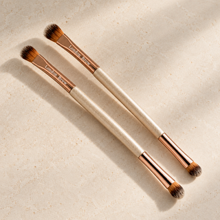 Two London Brow eyebrow makeup brushes with rose gold ferrules and cream handles, arranged diagonally on a textured beige surface with soft shadows.