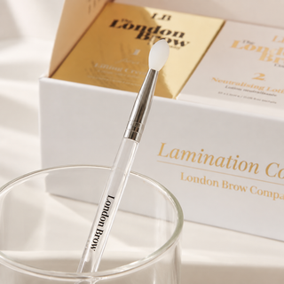 A clear cosmetic spatula applicator with a silver ferrule and white tip, branded London Brow, resting in a clear glass cup. In the background is the product packaging box with gold and white packaging and the London Brow brand name.