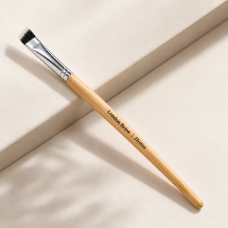 A flat-ended makeup brush with a light wood handle and silver ferrule, resting diagonally on a beige surface. The handle is printed with the text 'London Brow | Henna'.