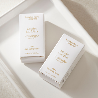 Two white cosmetic boxes from London Brow Company, one containing LashFixx Cystamine Lash Lift Ointment and the other Nourishing Lotion, arranged on a white tray.
