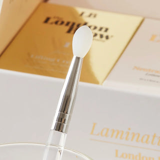 Cosmetic brush with a clear handle and white bristle tip on a white surface with 'London Brow' branding.