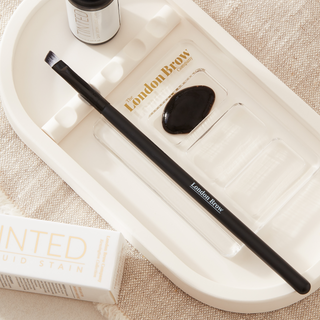 Eyebrow brush with black handle and white ferrule on a textured beige surface from the london brow company